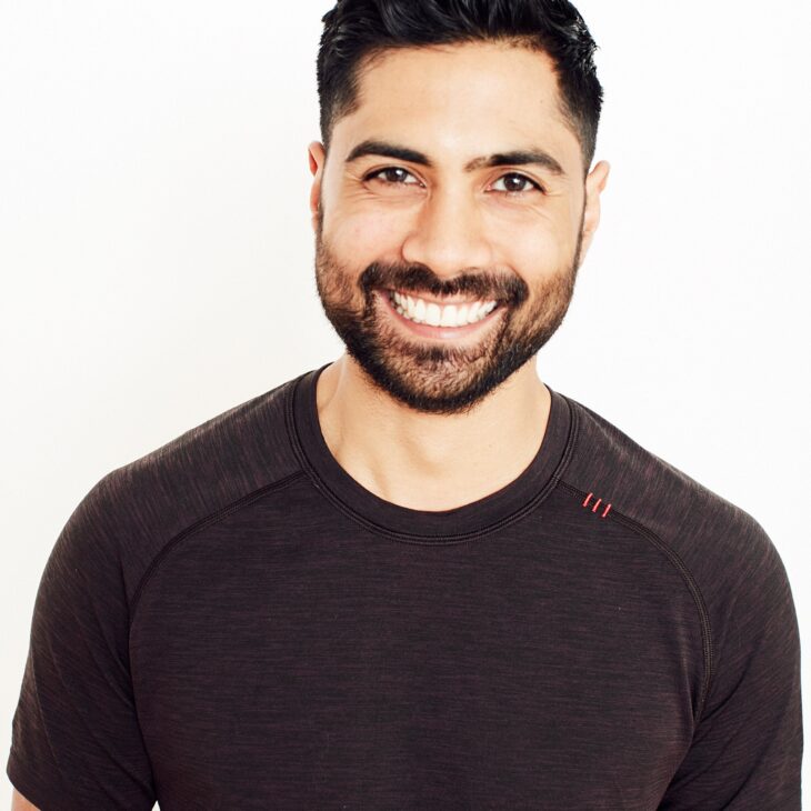 A person with dark short hair and dark facial hair and is wearing a brown t-shirt. They are smiling, facing forwards in front of a plain white background.