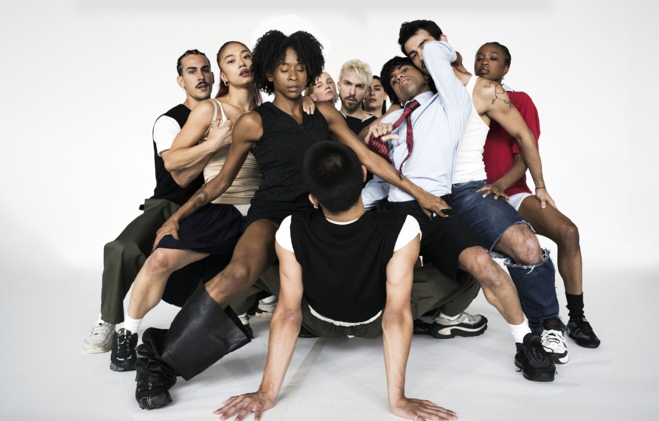 All dancers apart from one are in a deep second, overlapping each other facing inwards to the dancer on the floor. The dancers are holding on to each others legs with their arms for support. There is one dancer on the floor who is horizontal and supporting themselves with their arms fully extended to the floor, whilst looking at the dancers surrounding them.