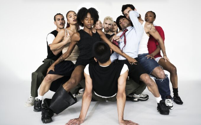 All dancers apart from one are in a deep second, overlapping each other facing inwards to the dancer on the floor. The dancers are holding on to each others legs with their arms for support. There is one dancer on the floor who is horizontal and supporting themselves with their arms fully extended to the floor, whilst looking at the dancers surrounding them.