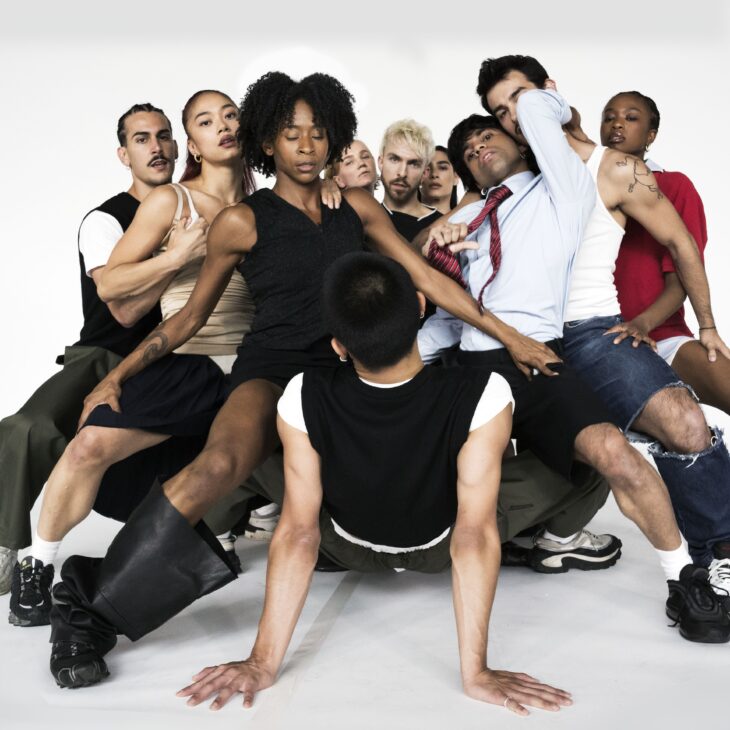 All dancers apart from one are in a deep second, overlapping each other facing inwards to the dancer on the floor. The dancers are holding on to each others legs with their arms for support. There is one dancer on the floor who is horizontal and supporting themselves with their arms fully extended to the floor, whilst looking at the dancers surrounding them.
