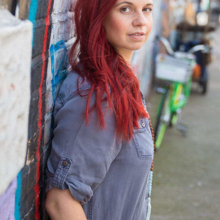 A woman who has bright red long hair and is wearing a denim blue shirt. She is on a street and her body is facing the right and she is looking towards the front with her head turned.