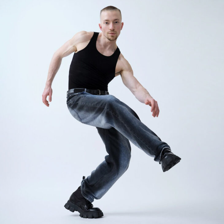A person with very short brown hair and is wearing a black vest top, blue jeans and black boots. He is balancing on his left leg that is bent and his right leg is bend and lifted up. His right arm is behind him whilst his left arm is out in-front reaching towards his right foot.