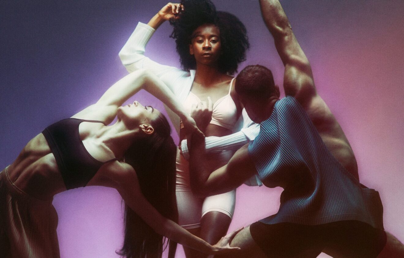 There are three dancers. The dancer on the left is creating a curve in their back with their arms extended holding on to the dancer in the centre who is holding their hair up with their arm and stepping forward. The dancer on the right is in a deep lunge towards the dancer in the centre with their left arm bent and their right arm extended upwards.