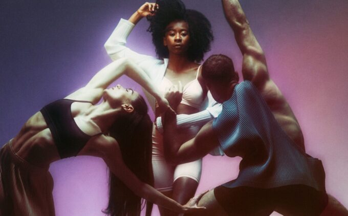 There are three dancers. The dancer on the left is creating a curve in their back with their arms extended holding on to the dancer in the centre who is holding their hair up with their arm and stepping forward. The dancer on the right is in a deep lunge towards the dancer in the centre with their left arm bent and their right arm extended upwards.