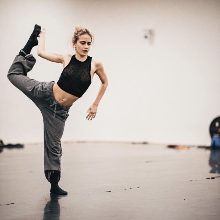 A dancer who has blonde hair which is tied up and is wearing a black sleeveless top, grey loose trousers and black socks. They are also in a balance on one leg whilst holding up their right leg back with their right arm, opening up their body to the front.