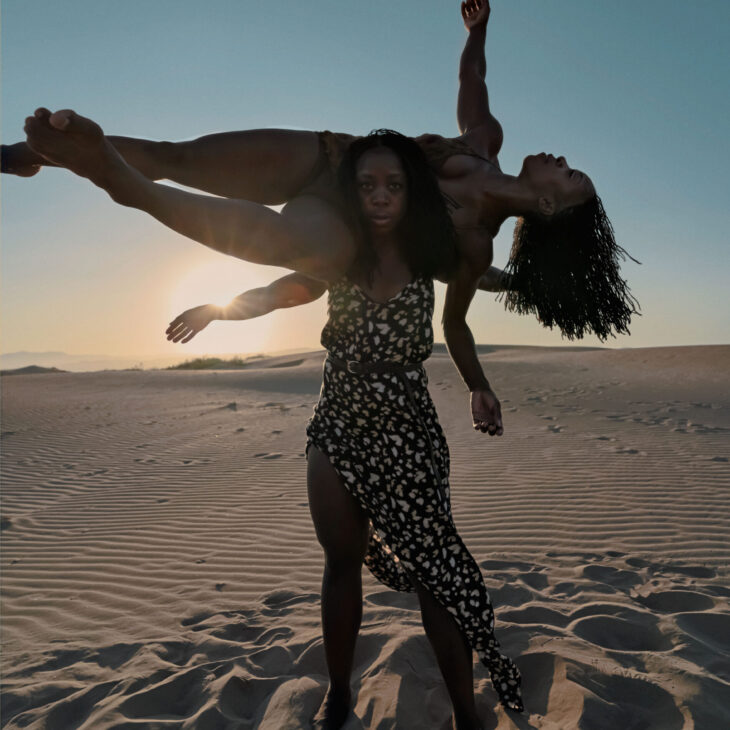 There are two dancers. One is standing vertical looking straight forward with their arms extended out to the side. The other dancer is horizontal, balancing on the other dancers shoulders with their arms extended out to the side whilst looking upwards towards the first one. They are on the sand with a sunset in the background.